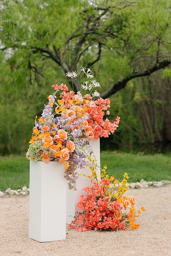 Wedding floral arrangement of roses and wildflowers on white pedestal plinths with trailing greenery on a green lawn near trees and rocks