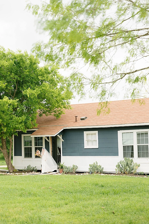Bridal portrait of a bride with veil holding a colorful bouquet on a porch, long cathedral veil flowing beside a house exterior and lawn
