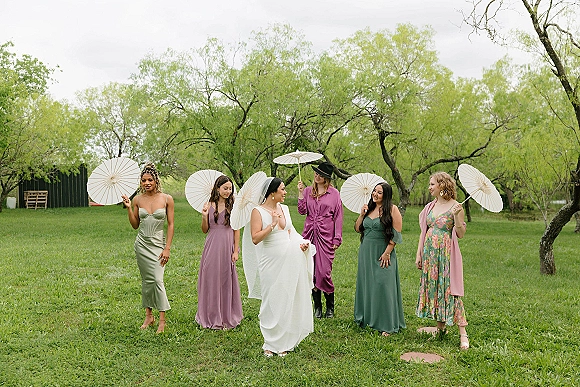 Bridesmaid group photo with bride with bridesmaids holding white paper parasols in a grassy field near a barn under overcast sky