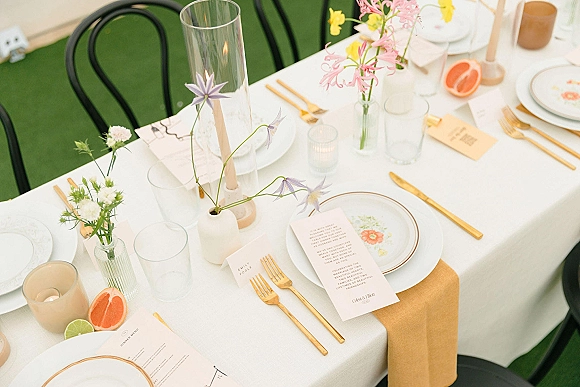 Reception tablescape with white and tan tablescape styling, gold flatware, floral plates, taper candles, pastel bud vases, and citrus on a lawn