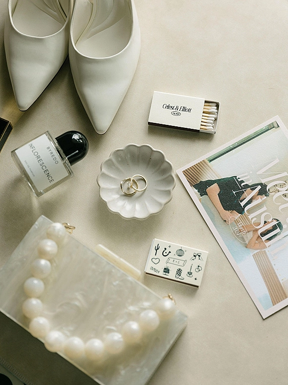 Wedding flat lay of white pointed-toe heels, gold wedding rings in a scalloped dish, pearl necklace, perfume, and stationery on light fabric