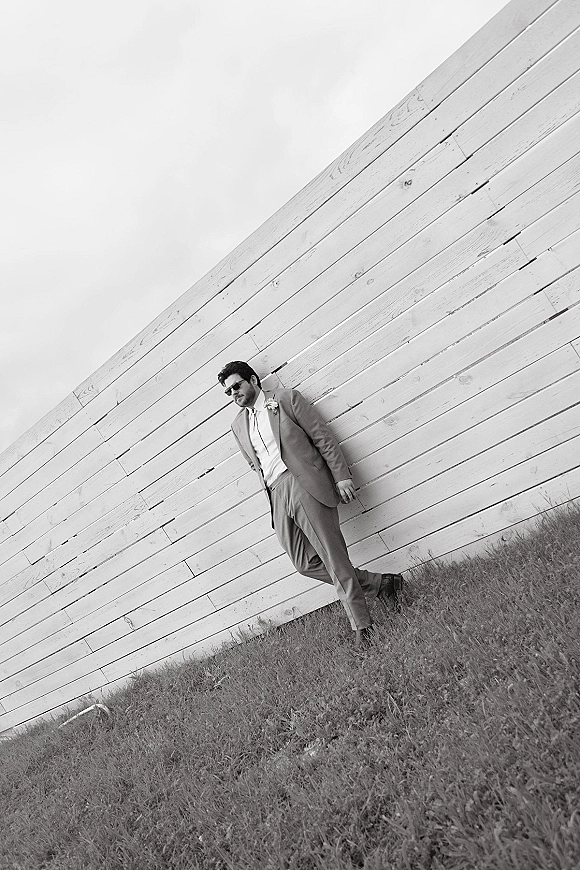 Groom portrait of a groom in sunglasses leaning against a wood plank wall, wearing suit and tie with a boutonniere outdoors