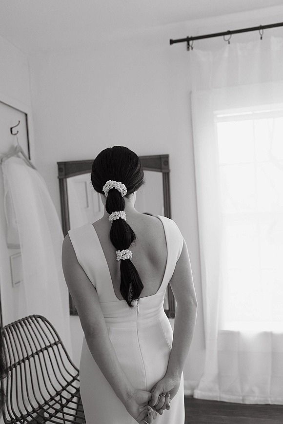 Bridal portrait of a bride in a backless wedding dress, pearl hair accessories and ponytail, looking in a mirror by window light