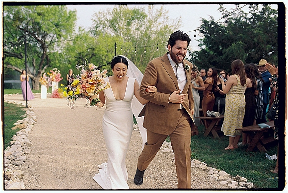 Wedding recessional as bride and groom walk the aisle, bride lifting a wildflower bouquet and veil, guests cheer under string lights in garden