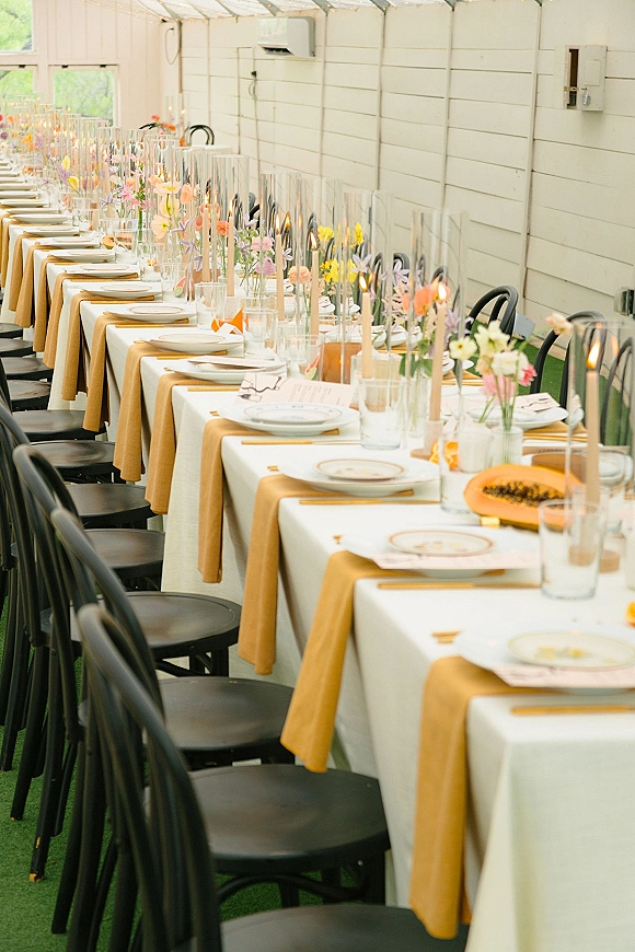 Reception tablescape with long banquet table wedding styling, mustard runners, bud vases, taper candles in glass hurricanes under a tent ceiling