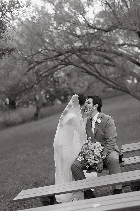 Wedding kiss portrait in black and white as the bride leans in, veil draped over them, bouquet on groom’s lap on a park picnic table