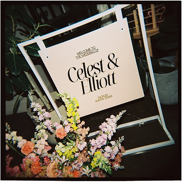 Wedding welcome sign in a white frame with peach-and-lavender floral arrangement, set against green foliage near indoor chairs
