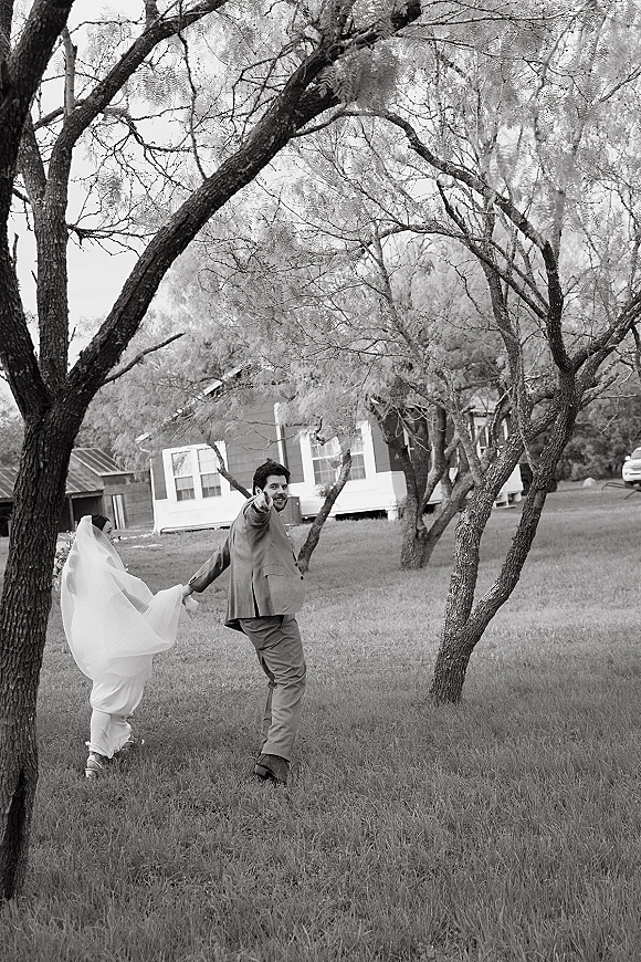 Wedding couple portrait of bride and groom walking away hand in hand, veil in motion as the groom looks back in a grassy field with trees