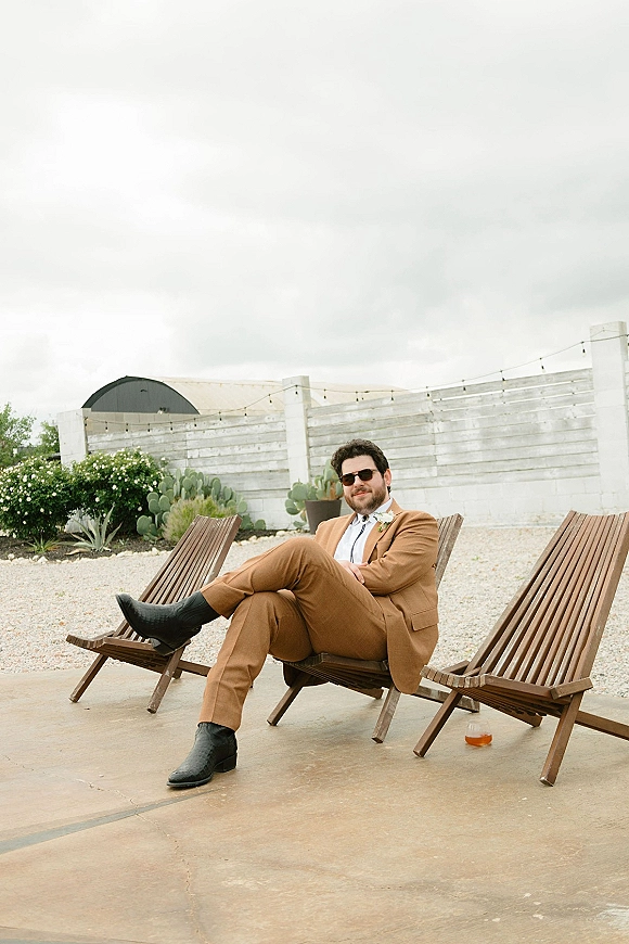 Groom portrait in a tan suit groom wearing sunglasses, seated in a wooden lounge chair on a cactus-lined patio under string lights