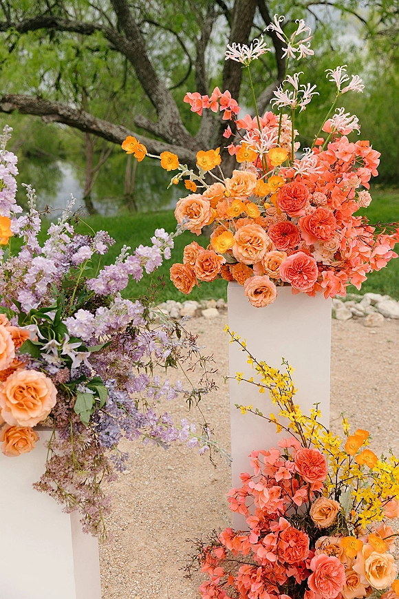 Wedding floral arrangements on white pedestal plinths with peach and coral roses, poppies, and greenery along a riverfront garden path