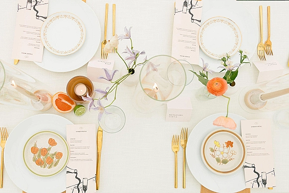 Wedding tablescape overhead with a white and gold table setting, patterned plates, menus, place cards, taper candles, bud vases, citrus accents