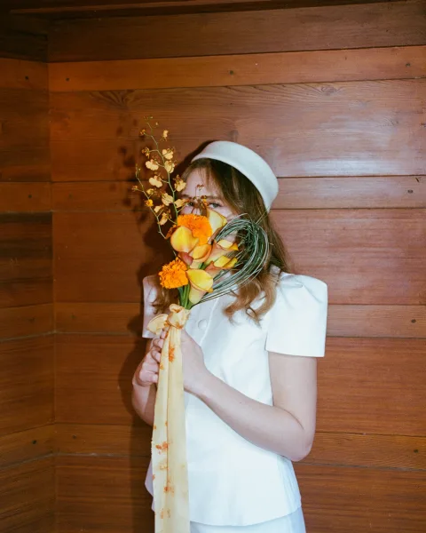 Bridal portrait of a bride holding bouquet covering her face, vibrant yellow and orange flowers with ribbon against a wood paneled wall