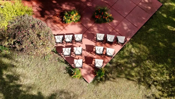 Ceremony seating layout with white folding chairs and floral arrangements in a semicircle on a lawn and tiled patio, dappled tree shadows