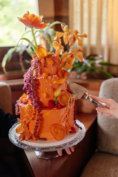 Wedding cake with orange wedding cake buttercream, orchids, citrus slices, berries, and sugar pearls on a glass stand by window light