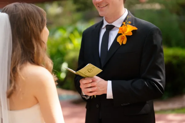 Wedding vows as groom reading vows from a booklet in black suit with orange orchid boutonniere beside veiled bride in garden courtyard