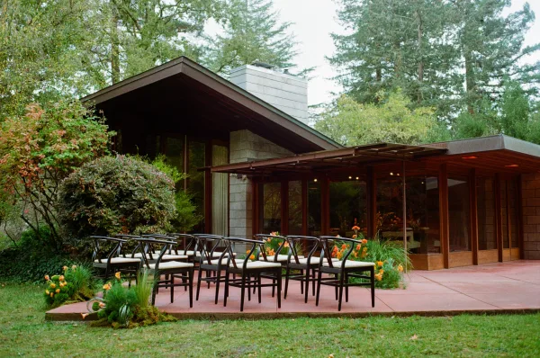 Ceremony setup for an outdoor wedding ceremony with patio chairs and greenery-lined aisle, orange florals, and a mid century modern house backdrop