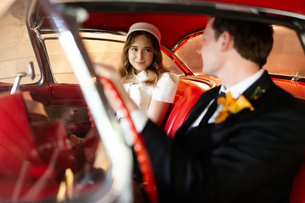 Couple portrait in a wedding car photo, bride in a pillbox hat and white dress gazing at groom in tuxedo on red leather seats