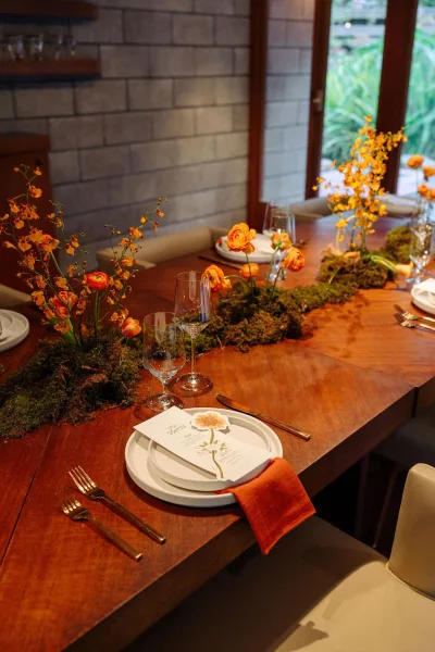 Reception tablescape on a wood wedding table with orange wedding centerpieces, moss runner, bud vases, and menus by brick wall windows
