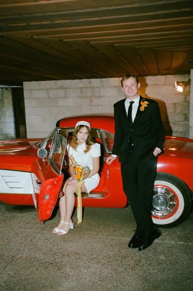 Couple portrait beside a vintage red car, bride in short dress and hat holding bouquet, groom in tuxedo by open door in garage