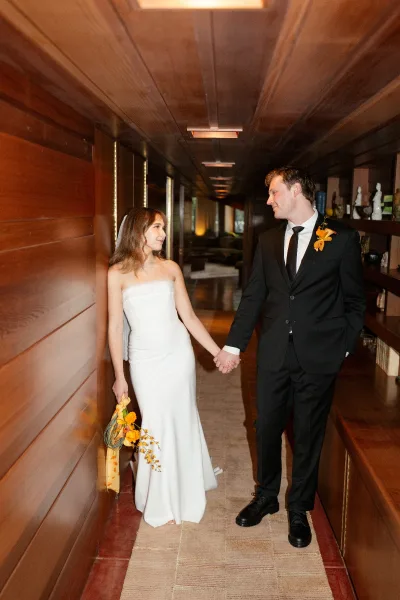 Couple portrait of bride and groom holding hands, walking down a wood-paneled hallway; she carries a bouquet with ribbon wrap