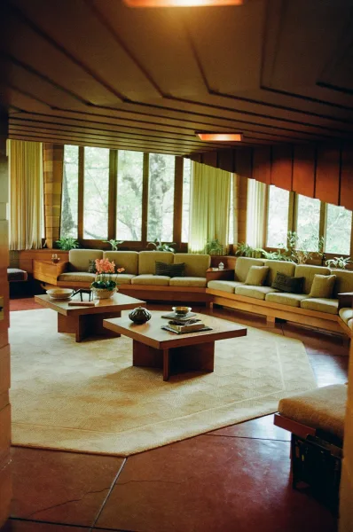 Mid century lounge with sunken living room seating, sectional sofa and low coffee tables beneath a wood paneled ceiling, forest view through large windows