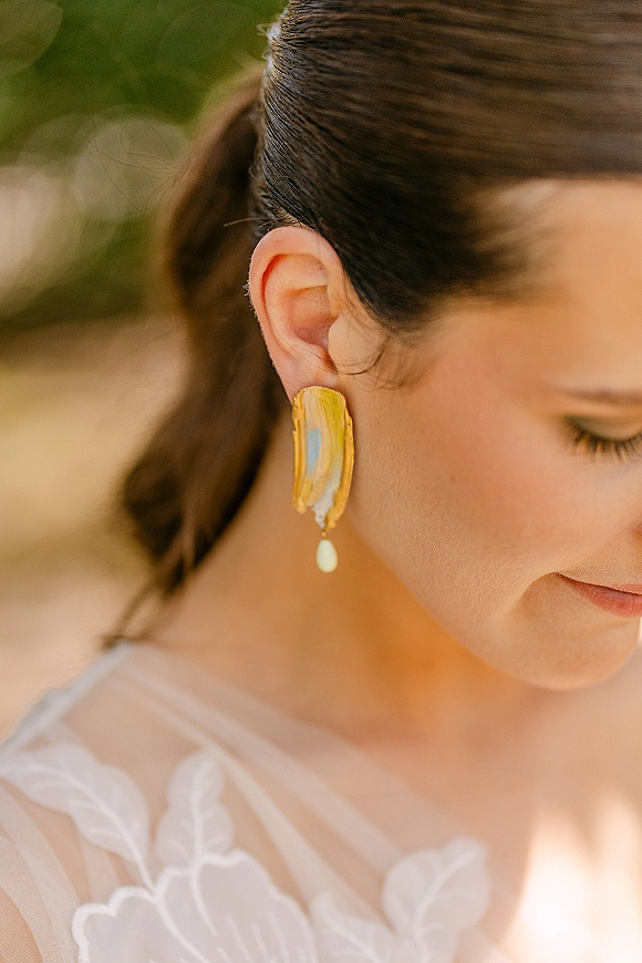 Bridal earrings in gold, statement bridal earrings hanging from a bride’s ear, with low ponytail and lace tulle gown against green bokeh outdoors
