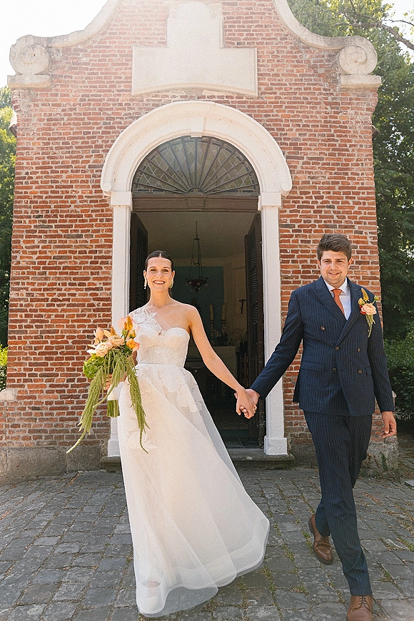 Couple portrait of bride and groom holding hands, her bouquet and his navy suit with orange tie outside a brick chapel doorway