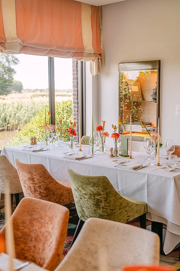 Reception tablescape with taper candle centerpieces, orange flowers and bud vases on a white tablecloth by a large window with countryside views