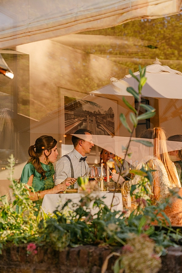 Wedding reception guests chat at a candlelit table with wine glasses and a floral greenery centerpiece by a restaurant window and patio umbrellas