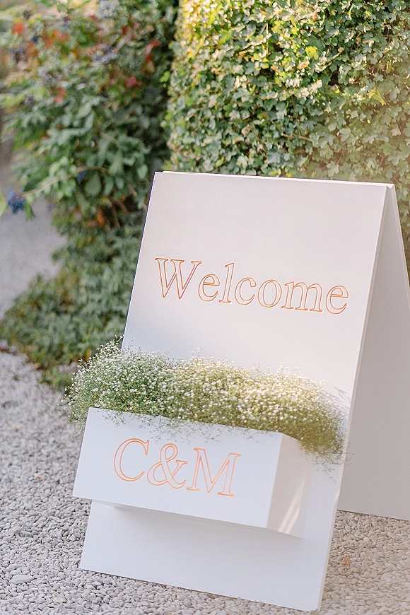 Wedding welcome sign with peach lettering and baby’s breath accent on a stand, set against a green ivy wall along an outdoor walkway