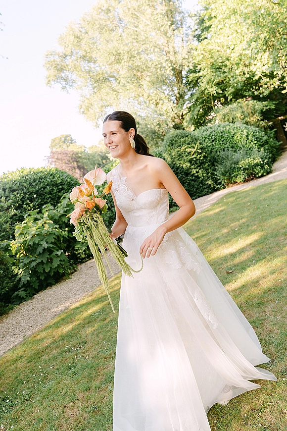 Bridal portrait of a smiling bride holding bouquet with anthurium, wearing a strapless lace gown on a sunlit garden path
