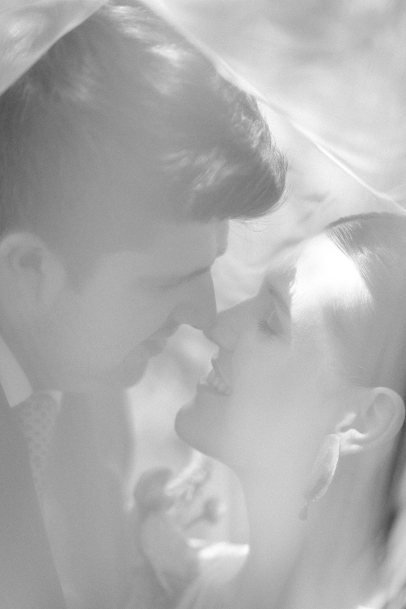 Wedding kiss portrait of bride and groom kissing under a bridal veil, her earrings visible, with backlit outdoor light and soft-focus foliage