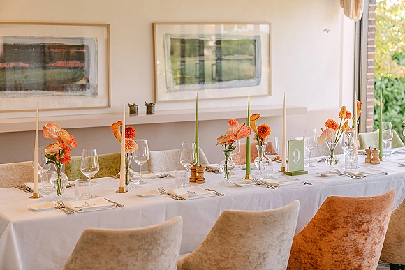 Reception tablescape with bud vase centerpieces of orange flowers and green taper candles on a white tablecloth, set by a window with framed art