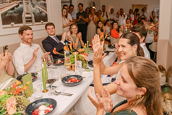 Wedding reception toast as guests applaud along a long banquet table with orange floral centerpieces, green taper candles, and glassware indoors