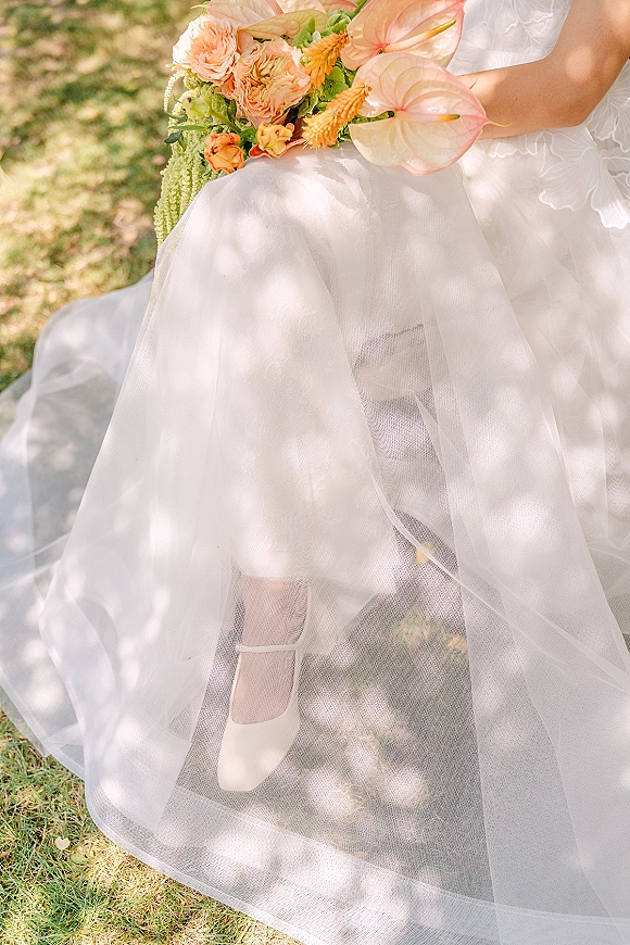 Bridal bouquet with peach anthurium bouquet blooms, orange roses and green foliage beside white tulle dress on a dappled grass lawn