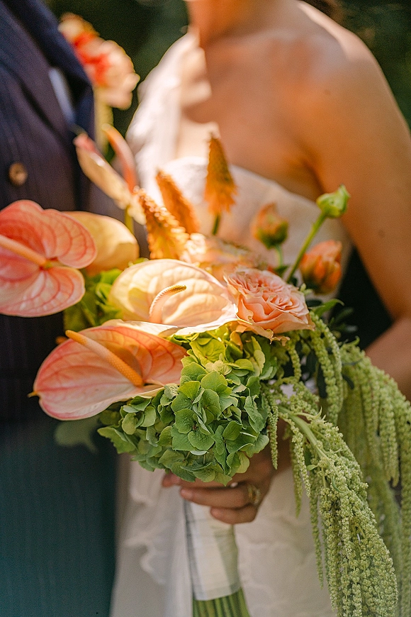 Bridal bouquet with anthurium bridal bouquet blooms, roses and hydrangea held by bride beside groom in suit, blurred garden behind