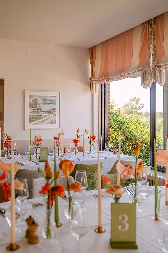 Reception tablescape with bud vase centerpieces, orange flowers, green taper candles, and white linens by a window with greenery view