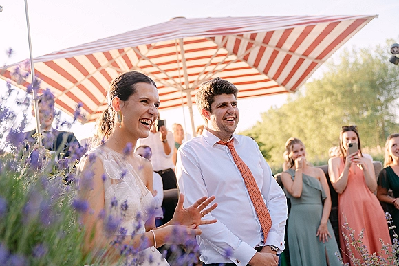 Reception speeches as the bride and groom laugh together, bride’s gown and groom’s orange tie under a striped patio umbrella in a garden