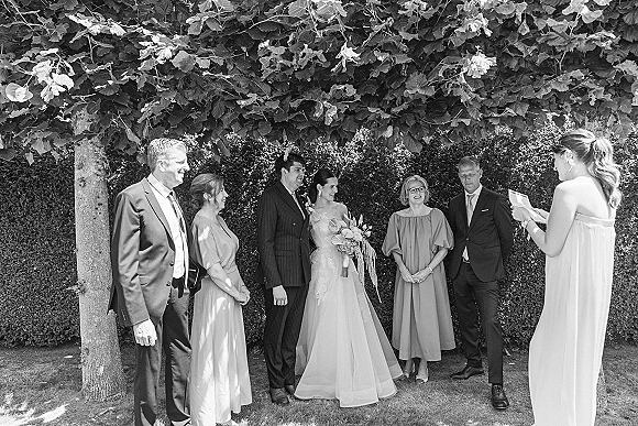 Wedding ceremony with couple exchanging vows as bride holds a cascading orchid bouquet, groom in dark suit, family watching by garden hedge