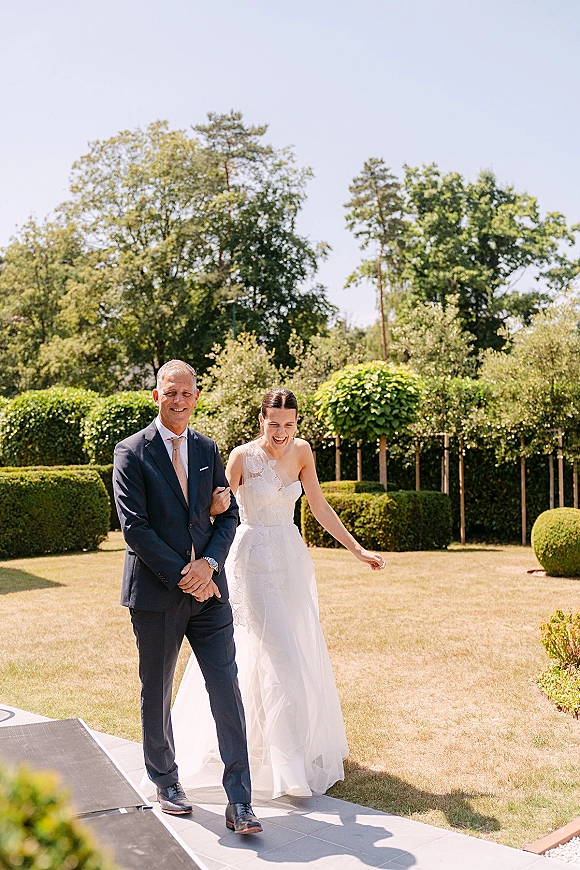 Processional moment as the bride walking down aisle smiles with her father on a stone garden walkway, dress and navy suit in bright light