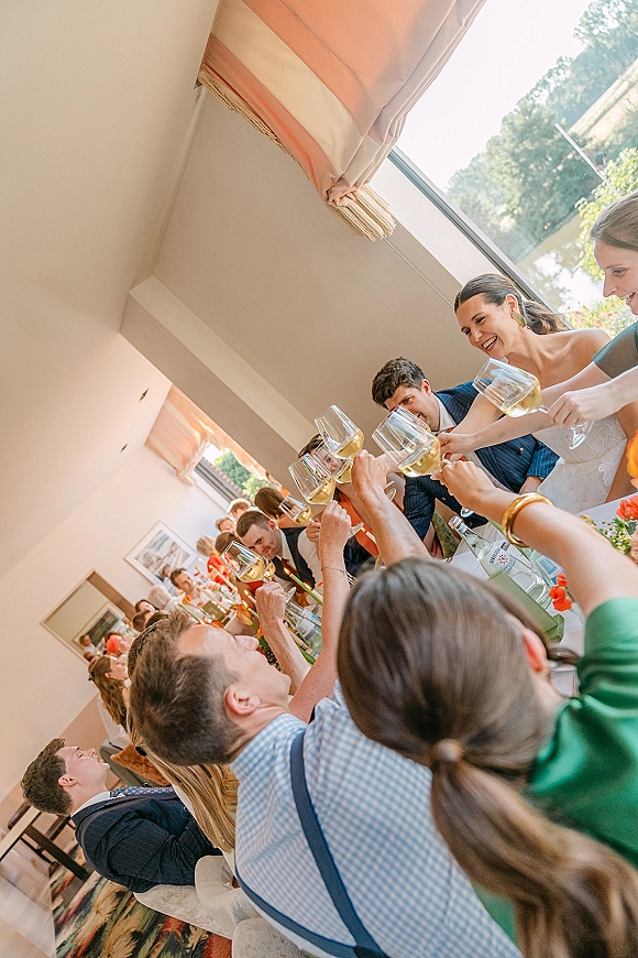 Wedding toast with guests raising champagne glasses over a long banquet table, taper candles and floral centerpiece in a bright windowlit reception