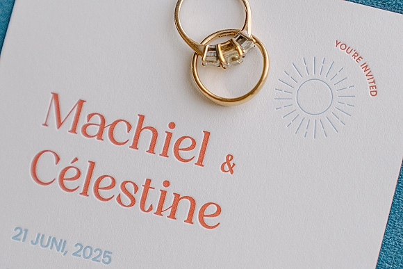 Wedding invitation suite with wedding rings flat lay, featuring gold bands and an emerald cut engagement ring on white paper over blue fabric