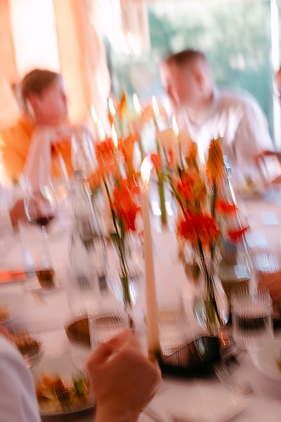 Reception tablescape with wedding table centerpieces in orange florals and taper candles, set with glasses and linens by bright windows and guests