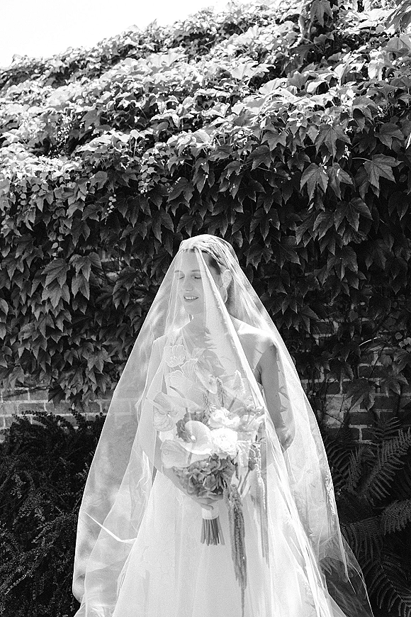 Bridal portrait in black and white of a bride wearing a veil, holding a calla lily bouquet beside an ivy-covered brick wall