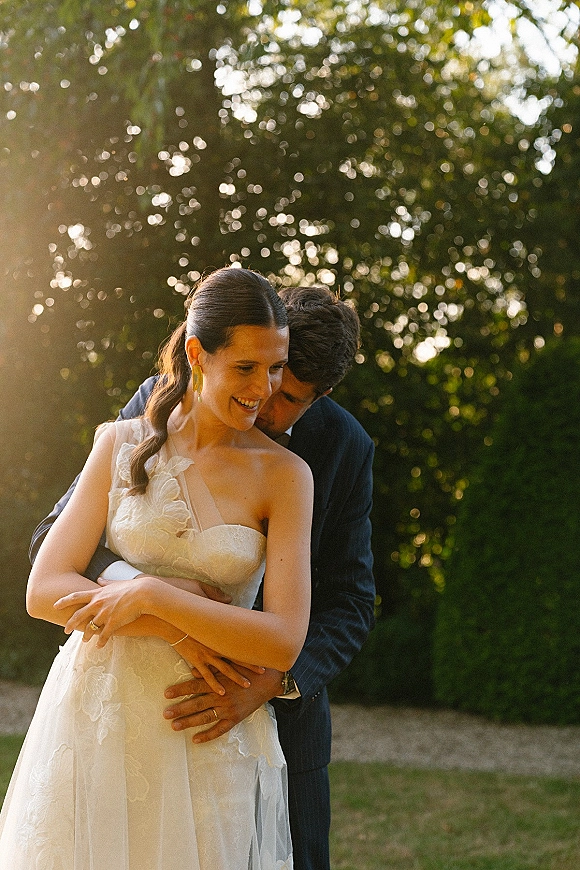 Couple portrait of bride and groom embrace as he hugs her from behind, her strapless lace dress glowing against sunlit garden trees