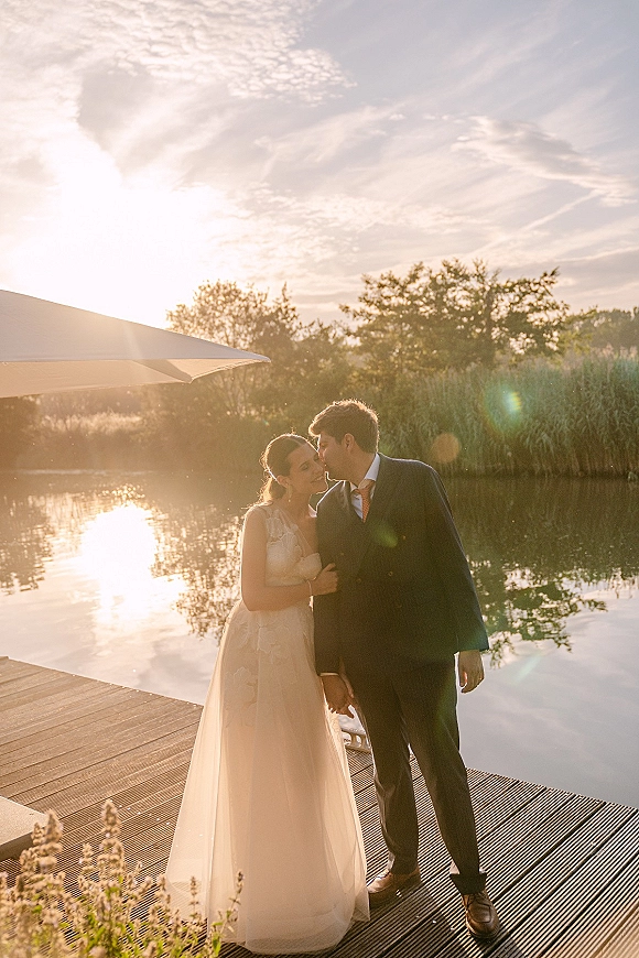 Wedding kiss portrait of bride and groom holding hands on a lakeside dock at sunset, bride in a lace bodice as sun flares behind them