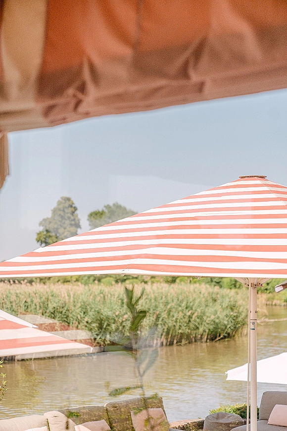 Outdoor lounge area wedding lounge seating with a striped patio umbrella over a sofa and pillows by a pond with reeds and trees