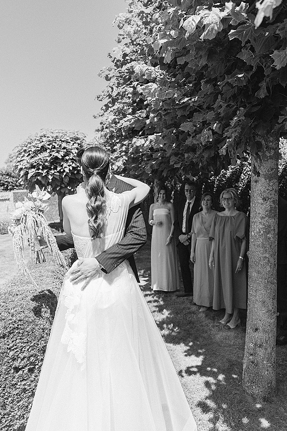 First look moment as bride and groom embrace, bride holding bouquet with long veil in a sunlit garden while bridesmaids watch
