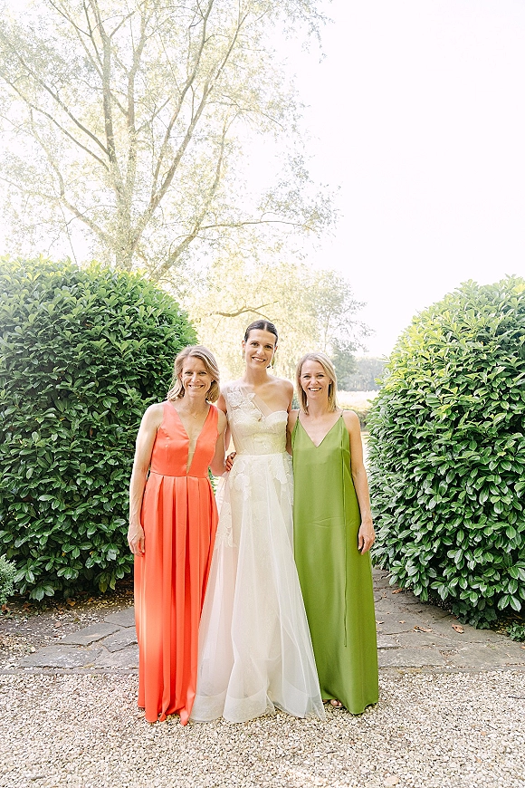 Bride with bridesmaids portrait showing the bride in a lace bodice gown beside coral and green dresses on a garden stone walkway in daylight