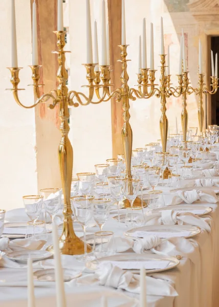 Reception tablescape with gold candelabra centerpieces, framed by stone columns and window openings against a plaster wall.